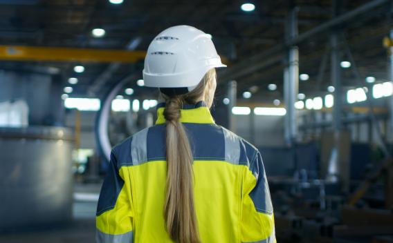 Shot of Female Industrial Worker in the Hard Hat Walking Through Heavy Industry Manufacturing Factory. In the Background Various Metalwork Project Parts 