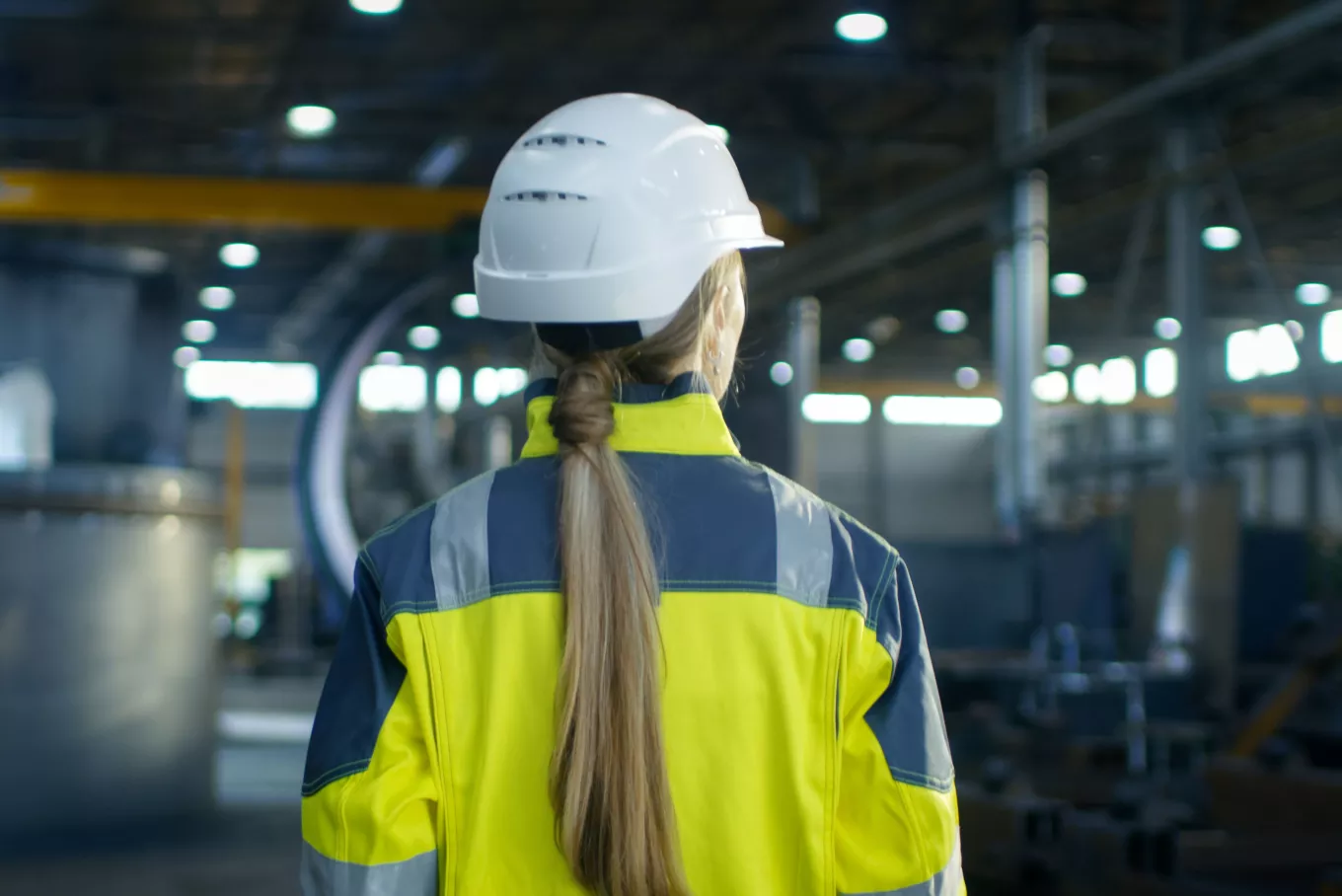 Shot of Female Industrial Worker in the Hard Hat Walking Through Heavy Industry Manufacturing Factory. In the Background Various Metalwork Project Parts 