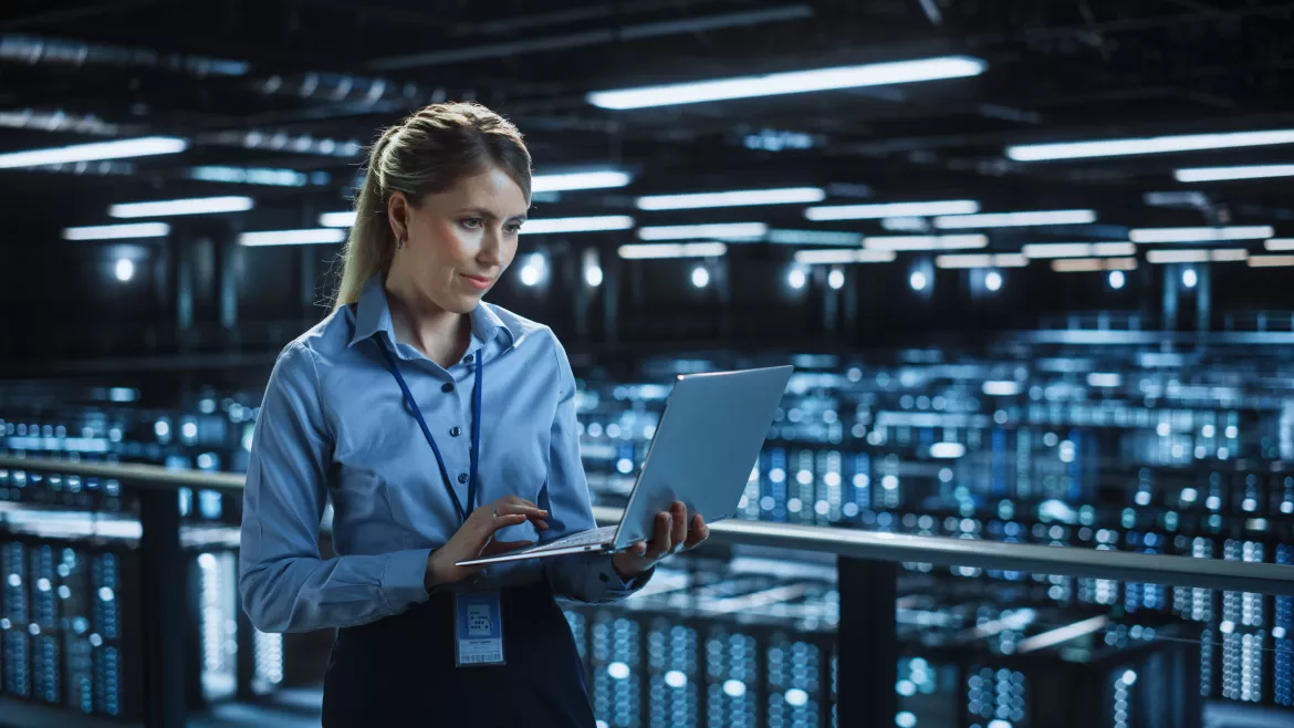 Portrait of IT Specialist Using Laptop in Data Center. Big Server Farm Cloud Computing Facility with Female System Administrator Monitoring. Cyber Security, Network Protection