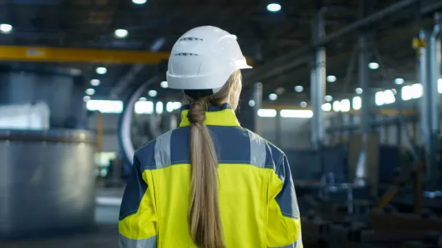 Shot of Female Industrial Worker in the Hard Hat Walking Through Heavy Industry Manufacturing Factory. In the Background Various Metalwork Project Parts 