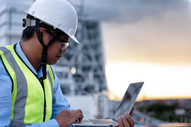 Civil Engineer wearing high visibility safety vest and helmet while using laptop looking to drawing detail on the terrace of decaying building at construction site. Surveyor for renovation concept