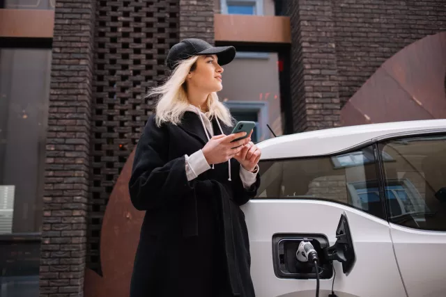 Using smartphone while waiting. Woman on the electric cars charge station at daytime. Brand new vehicle.