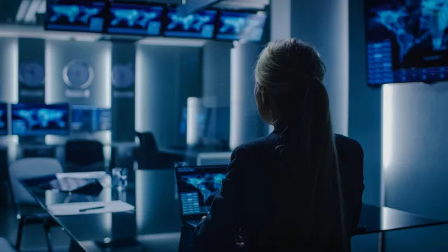 Female Special Agent Works on a Laptop in the Background Special Agent in Charge Talks To Military Man in Monitoring Room. In the Background Busy System Control Center with Monitors Showing Data .