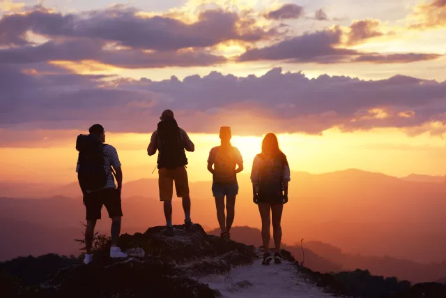 Group of four tourists or friends stands on mountain top and looks at sunset. Peoples travel concept